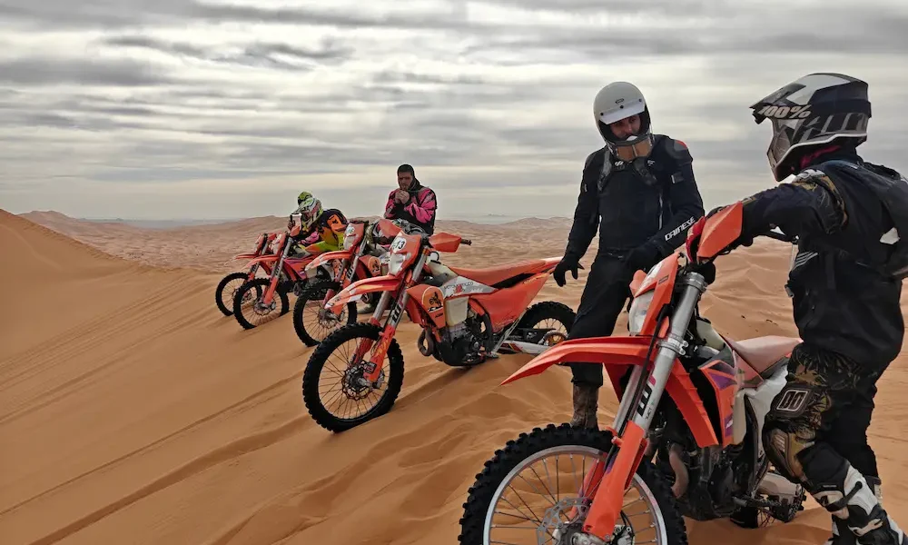 Pilotos de enduro de pie junto a su KTM ante las dunas de Merzouga